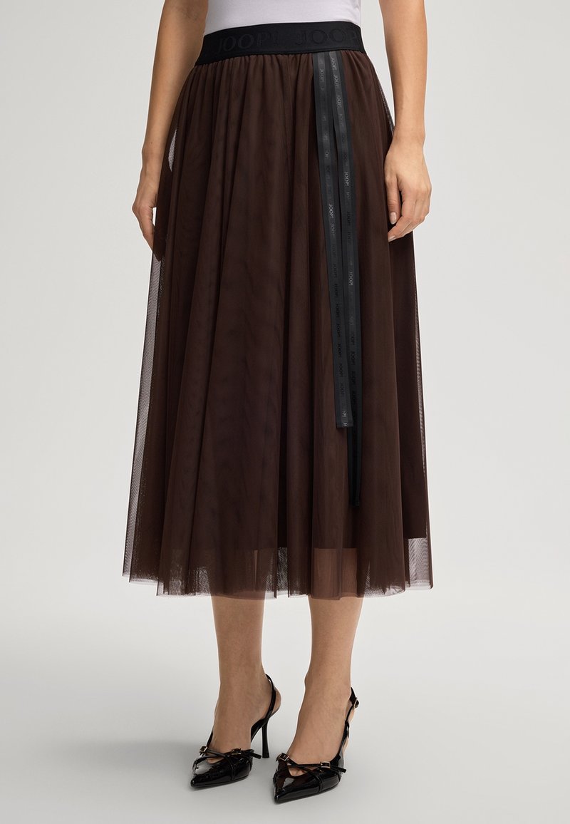 Femme portant une jupe midi en tulle marron avec une ceinture noire et un long ruban noir, associée à des chaussures à talons noirs.