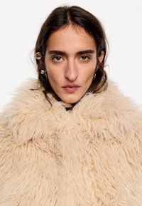Abrigo de pelaje beige con un alto cuello, que presenta un diseño texturizado y desordenado. El material parece suave y aterciopelado con una forma robusta.