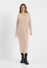 Beige gebreide jurk met een hoge halslijn, verlaagde schouders en geribbelde boorden. Heeft een rechte silhoet die tot halverwege de kuit reikt. Zwarte plateau laarzen.