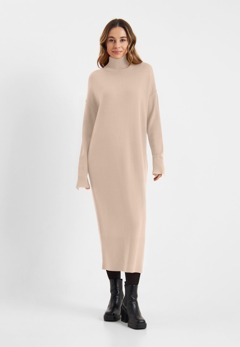 Beige gebreide jurk met een hoge halslijn, verlaagde schouders en geribbelde boorden. Heeft een rechte silhoet die tot halverwege de kuit reikt. Zwarte plateau laarzen.