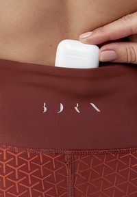 Bordowe legginsy sportowe z geometrycznym wzorem, z bezpieczną kieszenią na białe bezprzewodowe słuchawki oraz srebrnym nadrukiem logo.