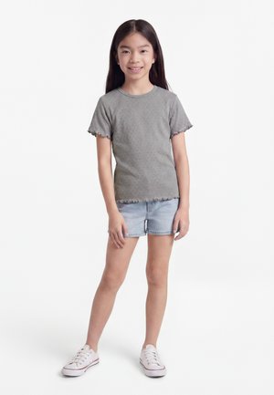 Mladé dievča s dlhými tmavými vlasmi, oblečené v sivom tričku s volánikovými rukávmi, svetlo rifľových šortkách a bielych teniskách, stojace a usmievajúce sa.