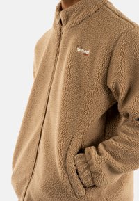 Giacca in pile beige con zip frontale, tessuto morbido e testurizzato, polsi elastici e logo ricamato "Schott NYC" sul petto.