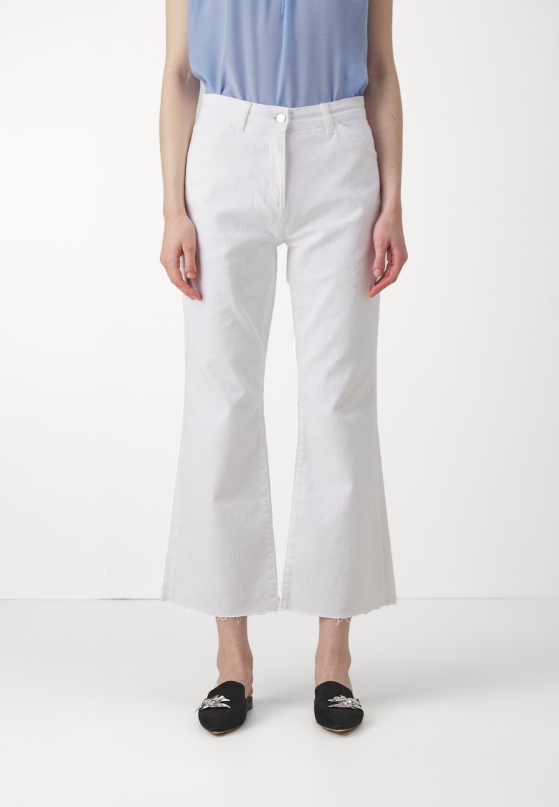 Jean flare blanc avec un ourlet effiloché, en tissu denim, associé à des chaussures noires à enfiler avec des accents décoratifs.