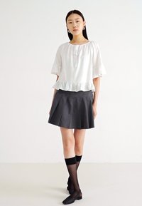 Blouse blanche à manches courtes avec un ourlet à volants ; associée à une jupe plissée sombre. Le modèle porte des chaussettes noires montantes aux genoux et des ballerines noires.