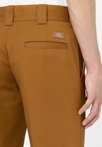 Bruna skräddarsydda byxor i en slät tygkvalitet, med bälteshällor, bakficka och en liten logopatch på höger sida.