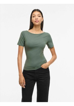 Frau mit langen dunklen Haaren, die ein eng anliegendes, olivgrünes Kurzarmshirt und schwarze Hose trägt, stehend mit einer Hand in der Tasche.