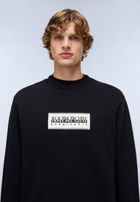 Black sweatshirt with a ribbed crew neckline, featuring a rectangular white logo patch with "NAPAPIJRI geographic" text in black and beige.