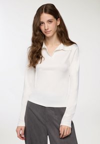 Maglietta polo bianca a maniche lunghe con colletto piatto, texture liscia, stile aderente e leggera lucentezza, abbinata a pantaloni grigi a gamba larga.
