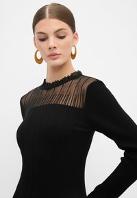Zwart geribd jurk met een transparant gestreept bovenlijfje en gerimpelde halslijn. Model draagt grote bruine oorbellen en heeft het haar netjes gestyled.