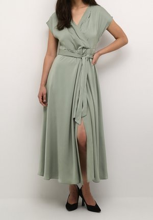 Robe longue vert clair style portefeuille, à manches courtes, taille nouée et fente à l'avant ; conçue en tissu lisse et fluide. Portée avec des talons noirs.