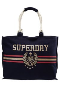 Sac fourre-tout bleu marine avec poignées en corde blanche, bandes dorées et rouges, et emblème "SUPERDRY ATHLETIC LEAGUE 50" avec étoiles et couronne de laurier.