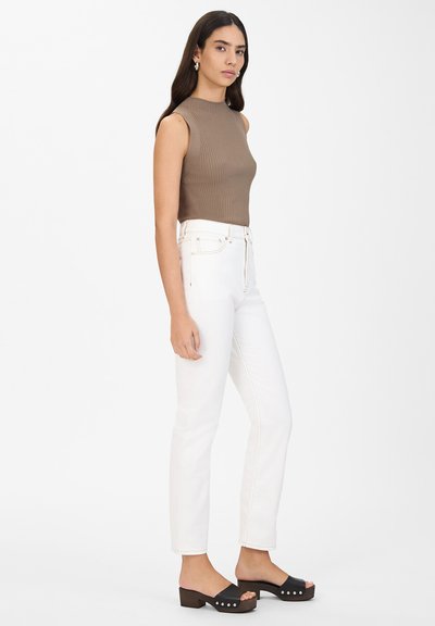 Top sin mangas de color marrón con relieve, jeans blancos de cintura alta y sandalias de plataforma negras con tachuelas, sobre un fondo blanco liso.