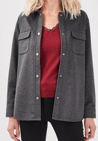 Szara koszula-kurtka z mieszanki wełny z dwiema kieszeniami, srebrnymi zatrzaskami i lekko teksturowanym wykończeniem, nałożona na czerwony top.