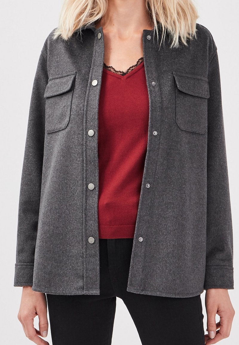 Szara koszula-kurtka z mieszanki wełny z dwiema kieszeniami, srebrnymi zatrzaskami i lekko teksturowanym wykończeniem, nałożona na czerwony top.
