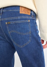 Personne portant un jean bleu Lee avec des coutures visibles sur la poche arrière et une étiquette en cuir de la marque, associé à un pull en maille crème.