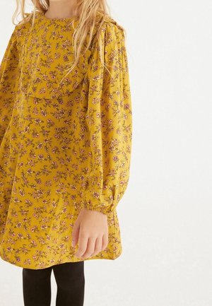 Abito giallo a fiori con lunghe maniche a sbuffo, caratterizzato da un scollatura arricciata e piccoli fiori rosa, indossato con leggings neri.