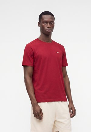 Homme portant un t-shirt rouge uni à manches courtes et un short beige clair, debout sur un fond blanc.