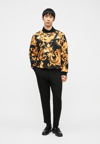 Νεαρός άνδρας που στέκεται με τα χέρια στις τσέπες, φορά μαύρο παντελόνι, μαύρα παπούτσια και μαύρο φούτερ με έντονα χρυσά σπειροειδή σχέδια.