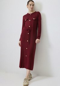 Pikk, burgundi värvi kudumdress, millel on pikad varrukad, kuldsed nööpide detailid, kaks rinnataskut ja vöökoht sidumise detailiga. Keskkleidi pikkus ja tekstureeritud kangas.