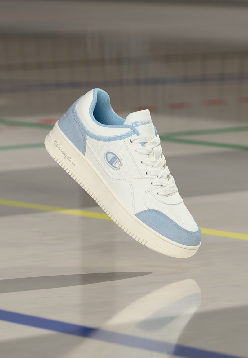 Λευκό και ανοιχτό μπλε αθλητικό παπούτσι Champion που αιωρείται πάνω από το πάτωμα ενός γυμναστηρίου με ορατές έγχρωμες γραμμές γηπέδου.