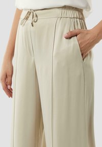 Lichtbeige losse broek met een elastische tailleband, trekkoordsluiting en zijzakken. Glad textuur met voorplooien.