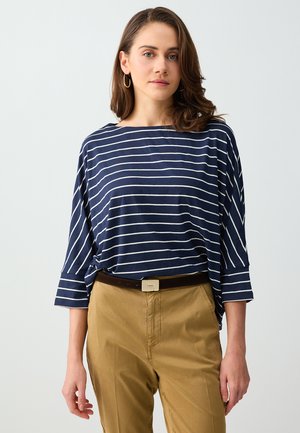 Kobieta z brązowymi włosami do ramion, nosząca złote kolczyki koła, granatowo-białą marynarską koszulę z długim rękawem oraz beżowe spodnie z ciemnym paskiem.