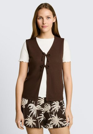 Vrouw draagt een wit shirt met korte mouwen, een bruin gebreid vest met strikjes aan de voorkant en bruine shorts met een beige palmboomprint.