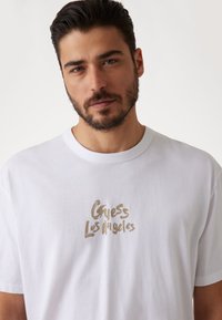 Hvid bomulds t-shirt med et guld, skrifttype "Guess Los Angeles" logo på forsiden, med et klassisk crew neck design og korte ærmer.