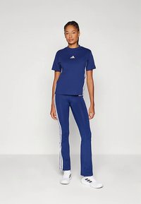 Γυναίκα που στέκεται φορώντας μια ναυτική μπλε φόρμα της Adidas με λευκές ρίγες και λευκά αθλητικά παπούτσια της Adidas, μπροστά από ένα απλό ανοιχτό φόντο.