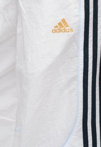 Vêtement de sport blanc à finition texturée, avec un logo "adidas" doré et des rayures verticales bleu foncé le long des côtés.