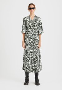 Rochie lungă, de tip wrap, cu un model abstract în verde și alb, având decolteu în formă de V, mâneci 3/4 și o fustă fluidă. Purtați cu ochelari de soare negri și cizme.