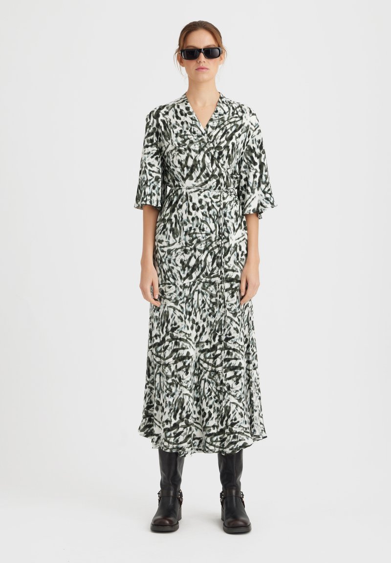 Rochie lungă, de tip wrap, cu un model abstract în verde și alb, având decolteu în formă de V, mâneci 3/4 și o fustă fluidă. Purtați cu ochelari de soare negri și cizme.