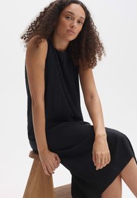 Black midi dress with pleated texture, sleeveless design, and side slit. Model seated on a wooden stool against a white background.