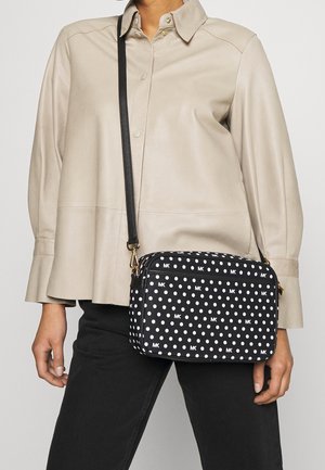 Zwarte tas met witte stippen en logo, rechthoekige vorm, verstelbare schouderband, gouden hardware accenten, geplaatst tegen een lichtbeige bovenstuk.