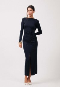 Femme portant une robe bleu marine à manches longues avec fronces sur les côtés et une fente à l'avant, accessoirisée avec des talons beige et des bracelets en or.