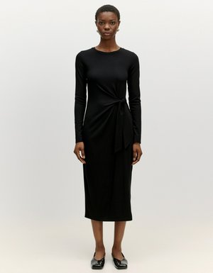 Black long-sleeve dress with a tied accent at the waist and a mid-length hem. Made from a smooth, stretchy fabric. Model wearing black flats.