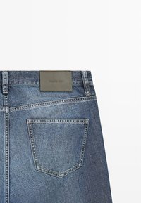 Zadní pohled na modré džínové kalhoty ukazující jednu kapsu a hnědý kožený štítek s názvem značky „Massimo Dutti“ na pásku.