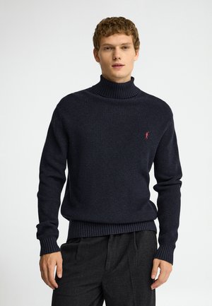Joven con un suéter de cuello alto acanalado azul marino oscuro con un pequeño logo rojo en el pecho y pantalones gris oscuro.