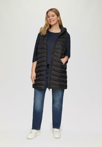 Glunderende vrouw in een zwarte donsjas over een marineblauw top, blauwe jeans en witte sneakers, staand met één hand in haar zak.