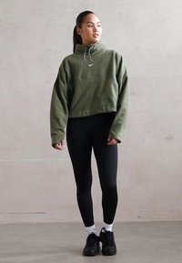 Olijfgroen cropped sweatshirt met hoge kraag en trekkoorden, gecombineerd met zwarte leggings en zwarte sneakers. Lichtgewicht materiaal.