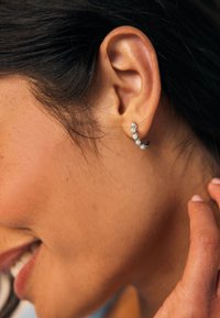 Gebogenes silbernes Creolen-Ohring mit kleinen, runden, klaren Edelsteinen. Glatte Metalloberfläche, schlankes Design und präzise Details in den Edelsteinen.