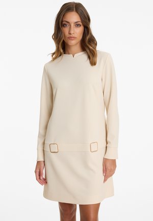 Robe beige à manches longues avec un léger col en V, mettant en avant deux accents en métal doré carrés à la taille et une texture lisse.