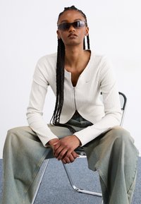 White ribbed zip-up top with long sleeves, paired with high-waisted light blue wide-leg jeans. Model wearing dark sunglasses, seated on a chair.