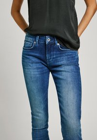 Jean en denim bleu avec un lavage usé, design à cinq poches et fermeture à bouton. Porté avec un haut noir ample.