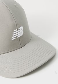 Hellgraue Baseballmütze mit gebogenem Schild und weißem gesticktem stilisierten "NB"-Logo auf der Vorderseite.