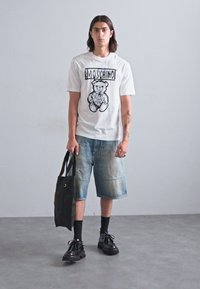 Tricou alb cu un grafic de urs negru și textul "MOSCHINO", purtat cu șorturi din denim albastre, șosete negre și încălțăminte neagră, ținând o geantă mare neagră.