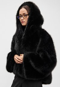 Juoda dirbtinio kailio striukė su gobtuvu, plačiomis rankovėmis, storu audiniu ir trumpu siluetu, dėvima ant juodų drabužių, rankos padėtos priekyje.