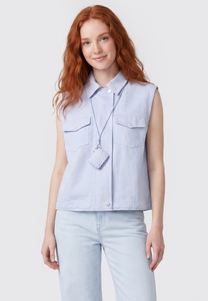 Junge Frau mit langen roten Haaren trägt ein hellblaues ärmelloses Shirt und hellblaue Jeans vor einem schlichten Hintergrund.