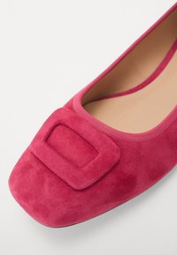 Rosa mokkaskinn leppesko med rund tå og en fremtredende firkantet spenne. Glatt tekstur med en bred elastisk kant ved åpningen.